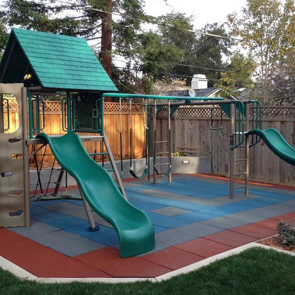 Backyard playground with Safe-Play Tiles in blue, gray, and terra cotta under a play structure with slides and climbing