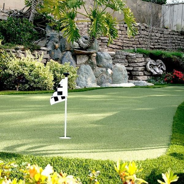 Backyard putting green with golf green artificial grass, featuring two flagsticks and surrounded by landscaping