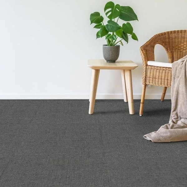 Foss Ribbed Carpet Tile — Smoke, installed in a living space with wicker chair and wooden side table