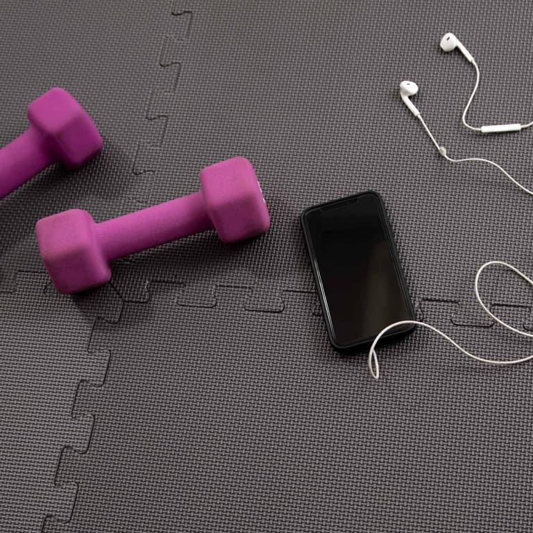 Gray fitness foam tiles with interlocking edges, shown under purple dumbbells, smartphone, and white earphones in a home